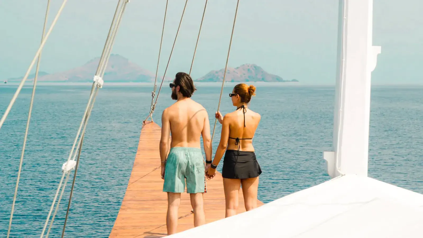 Couple holding hands at the bowsprit of a sailing boat, looking out at Komodo island hills
