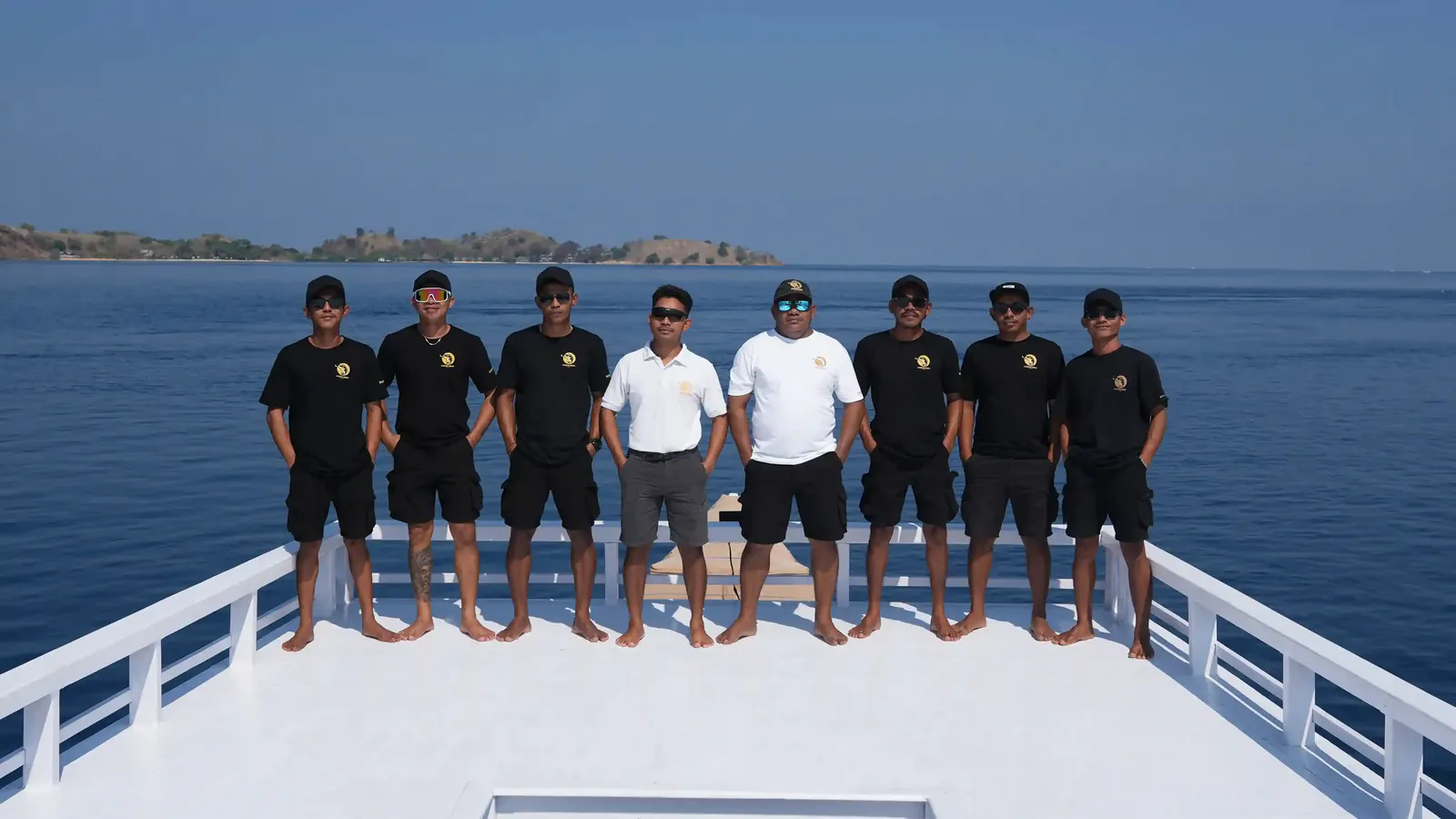 Eight boat crew members in matching uniforms and caps standing on a white bow with islands behind