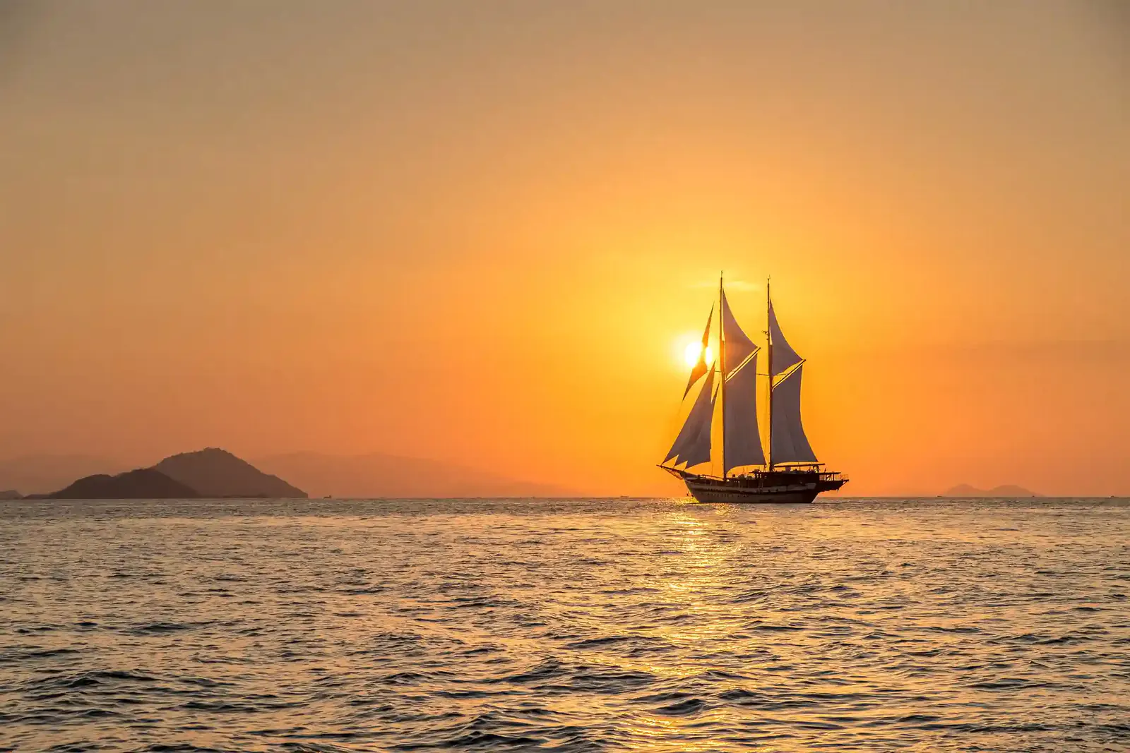 Two-masted sailing ship silhouetted against an orange sunset over calm sea near Komodo