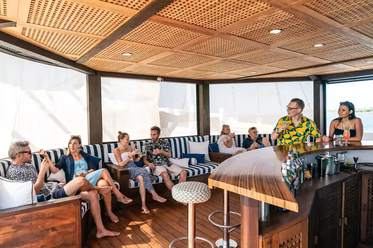 Guests chatting with drinks around the bar and lounge seating on a liveaboard's covered deck