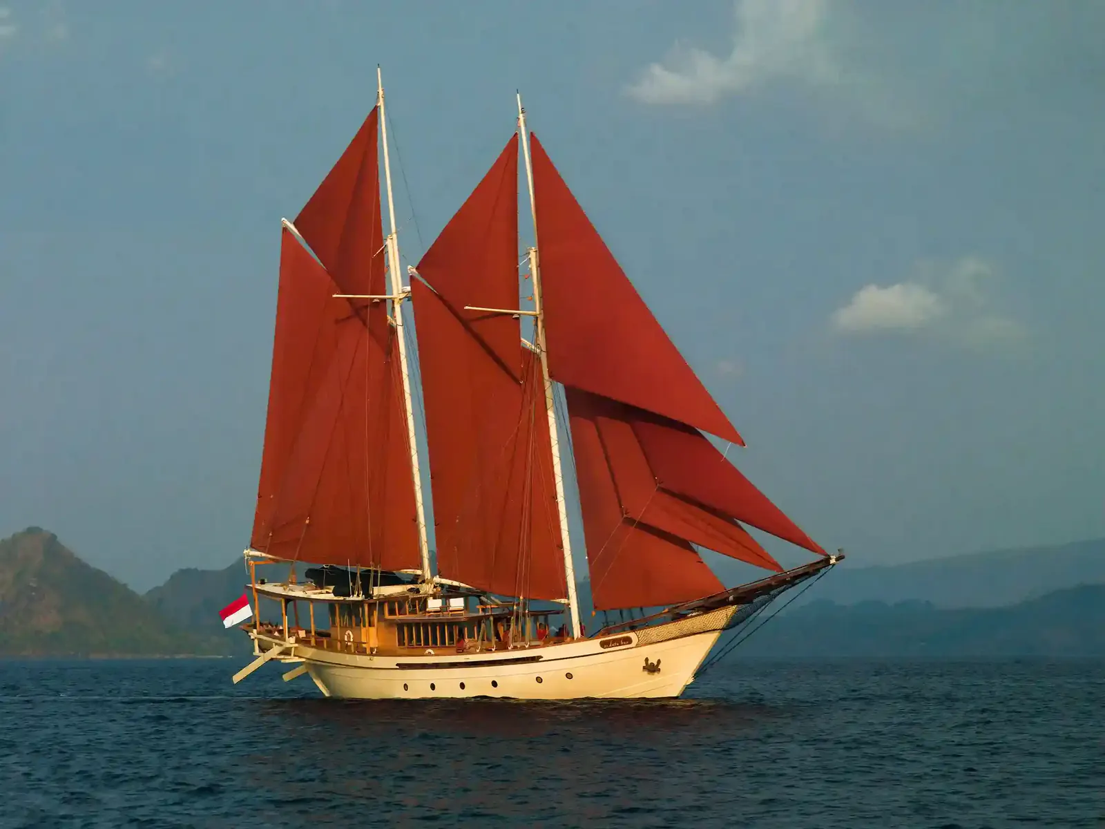 Luxury wooden schooner with red sails cruising past hazy island hills