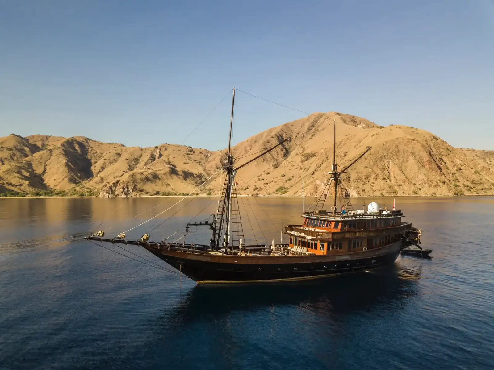 Dark-hulled wooden phinisi at anchor in golden light below dry hills in a calm Komodo bay