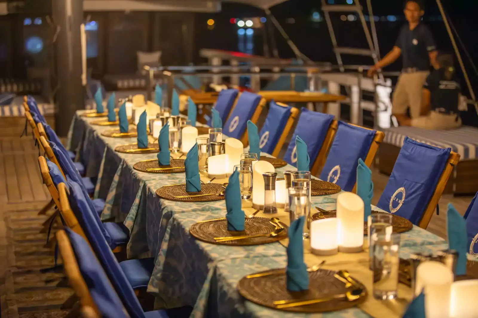 Candlelit dinner table set on a phinisi deck at night with blue chairs and a crew member nearby