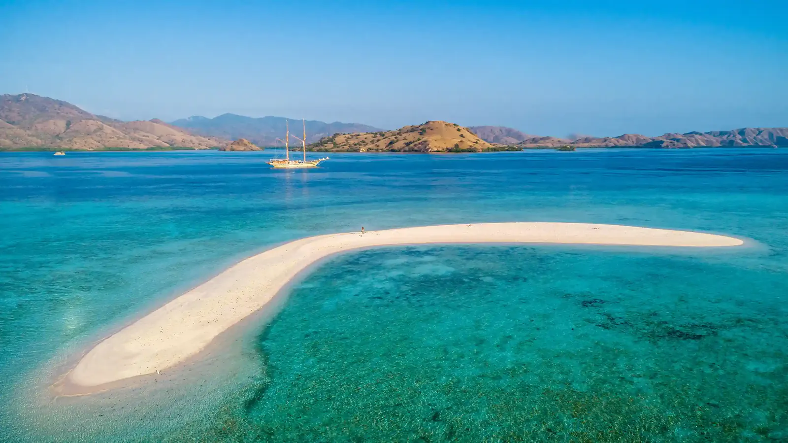 Curved white sandbar in turquoise shallows with a phinisi and island hills behind, Komodo